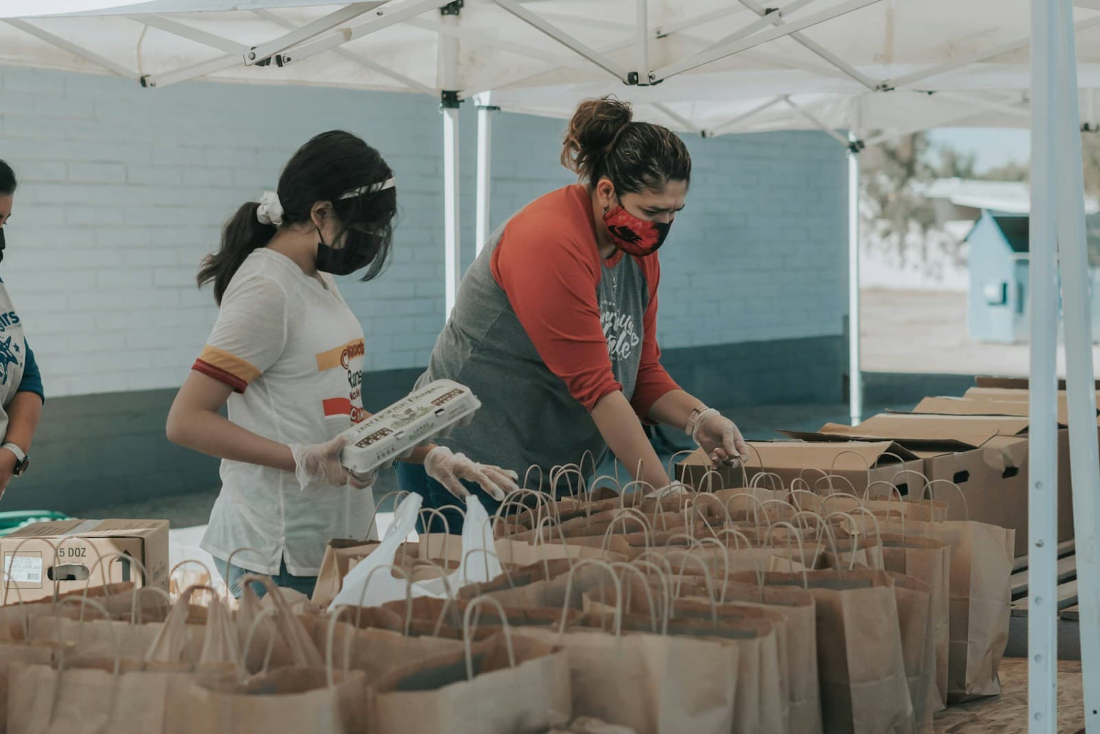 Two volunteers packing paper bags of food at a long table under a white canopy